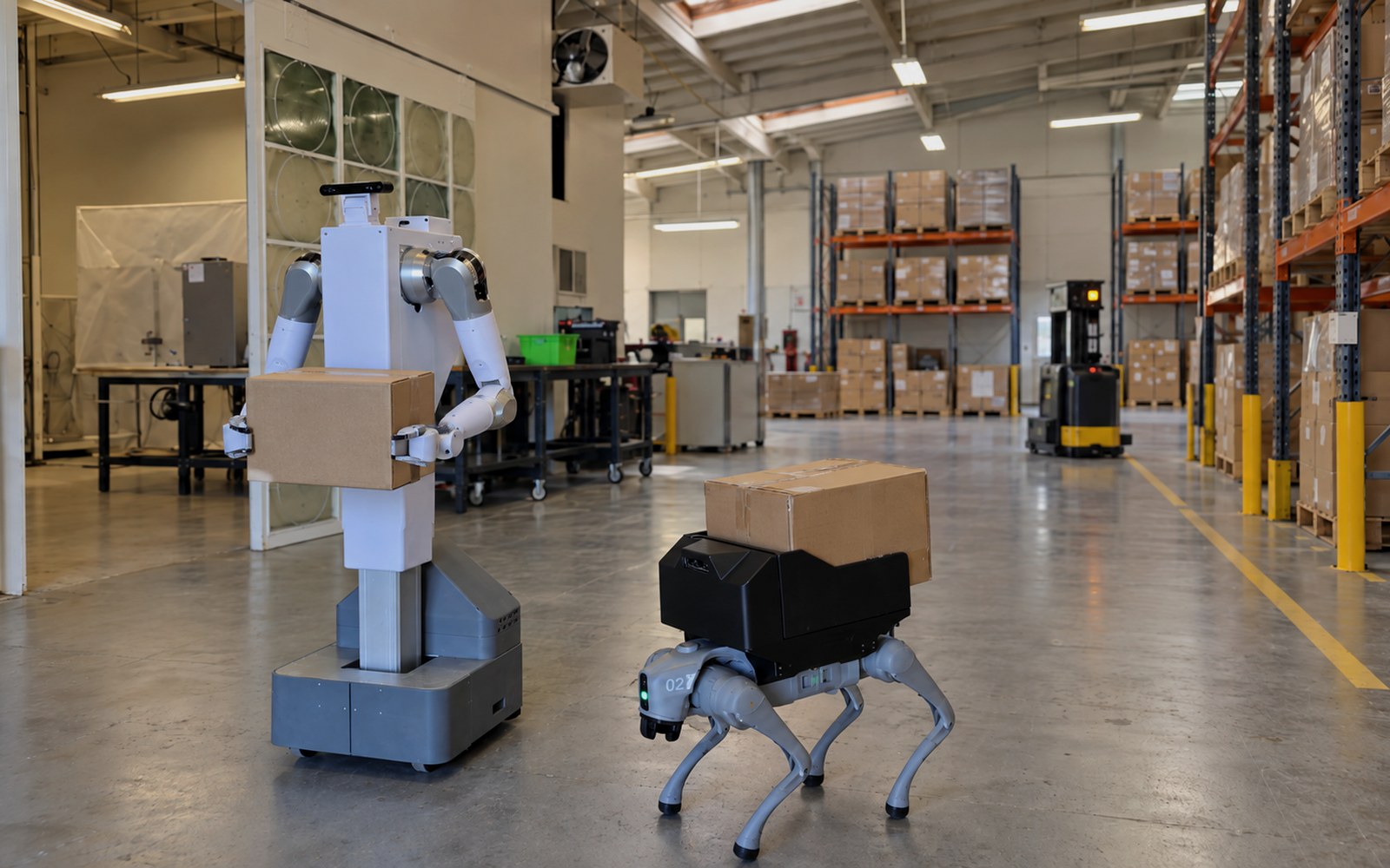 A mobile humanoid robot and a four-legged robot each carrying a parcel across the floor of a working warehouse alongside an autonomous forklift